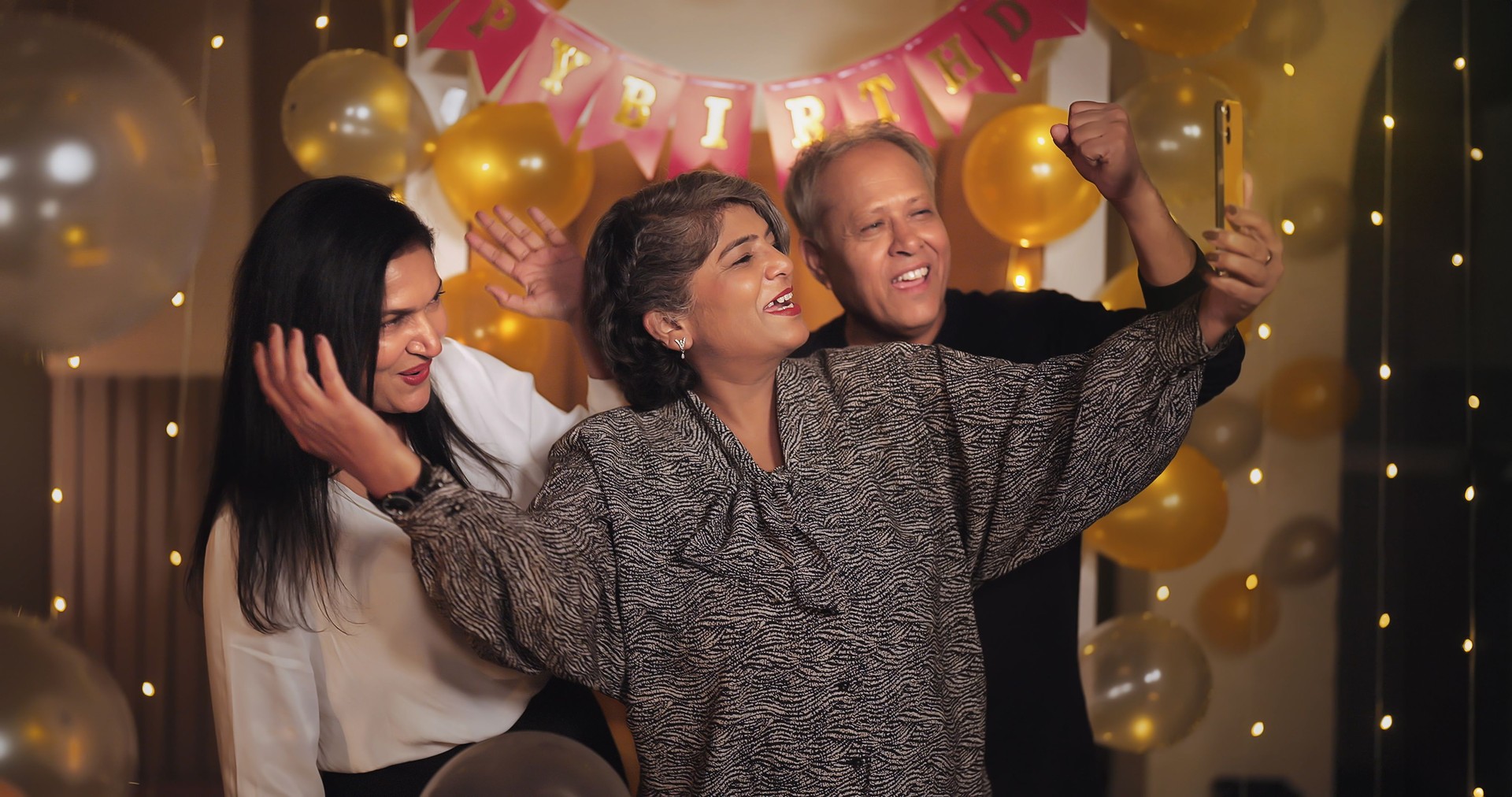 Indian smiling older aged cute male lady female friends hold cellphone take funny selfie night party indoor decor spend time house. Happy elder content creator family group having fun joy celebrate birthday enjoy together home